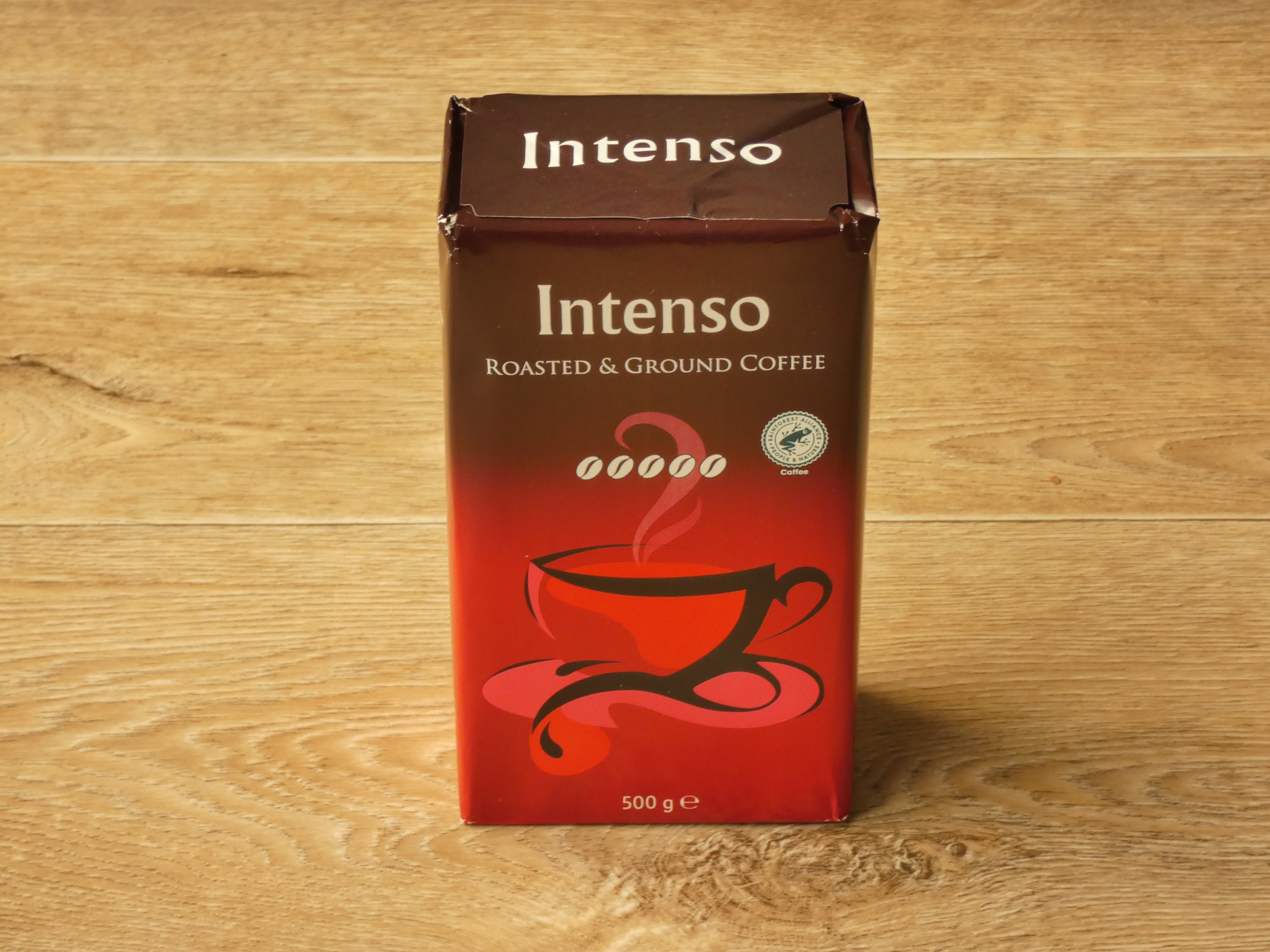 Dieses große Bild zeigt den Intenso Kaffee von Lidl. Es ist eine Vorderansicht der Verpackung. Die Verpackung ist oben braun und unten rot. Sie hat ‚Roasted & Ground Coffee‘ und ‚Intenso‘ im weißen Großbuchstaben und eine künstlerische Skizze von einer Kaffeetasse. Die Verpackung macht einen modernen und einfachen Eindruck.