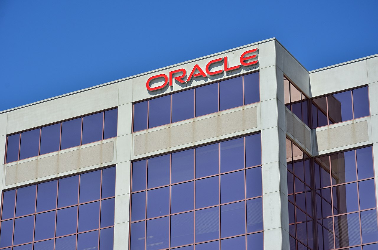 This large picture shows part of the Oracle building in Markham, Canada. We look up to a part of the outside of the building that has the company name in red letters on it. The building is made of clean raw concrete, and has mirroring windows. They are all closed and do not have windowsills. The sky is plain blue and the windows are blue as a result. The name of the company is at a top corner of the building, and there is yet another corner not far around that corner. We see that the windows follow regular patterns. The picture has a beautiful contrast between the red of the letters and the blue of the sky and the windows. Apart from that, the picture is very dull.